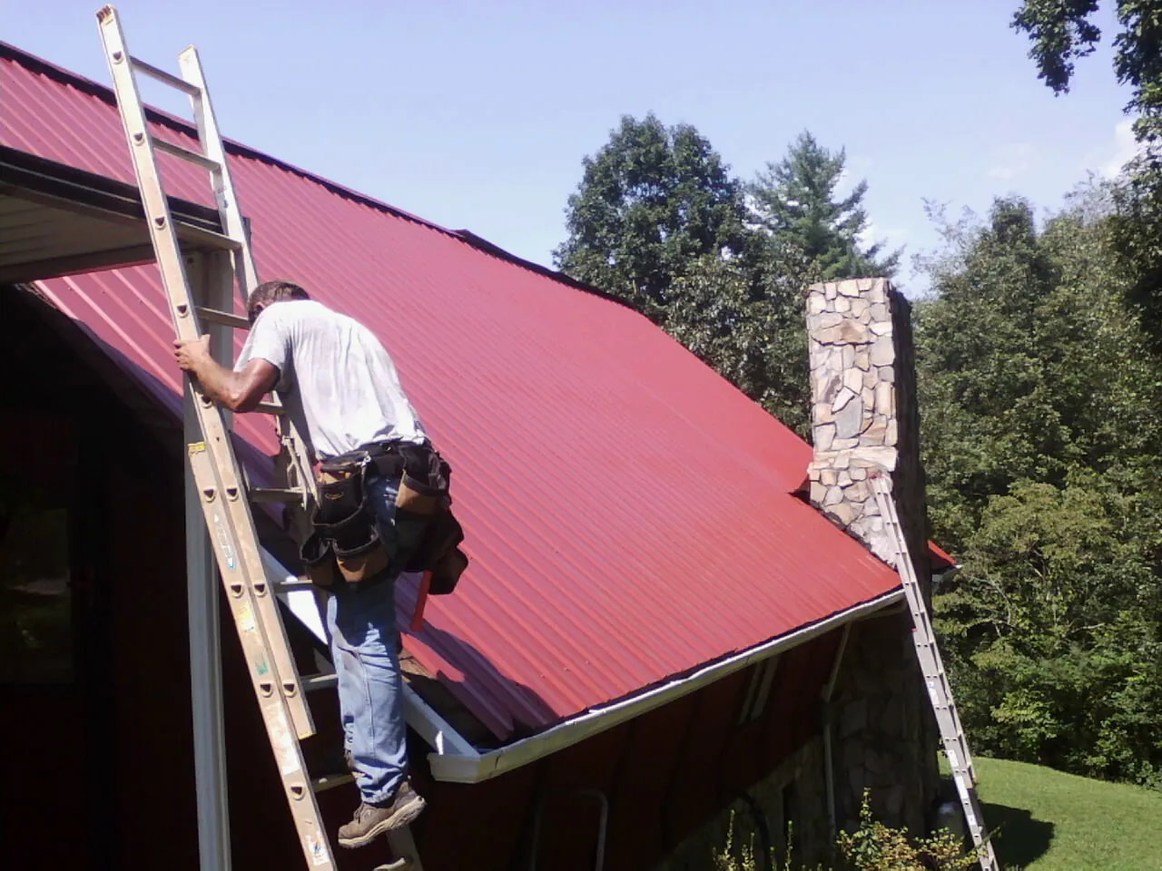 Roofer on a ladder beside a red metal roof and stone chimney — Soffit & Fascia Repair by Roofing Brothers in Rewey